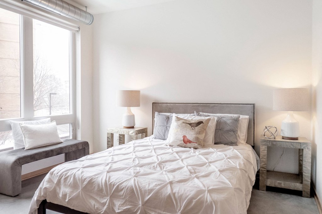 Bright bedroom here at 7west Apartment Homes with a neatly made bed, featuring white and gray linens and a decorative bird pillow. Soft lighting from two lamps creates a cozy ambiance.