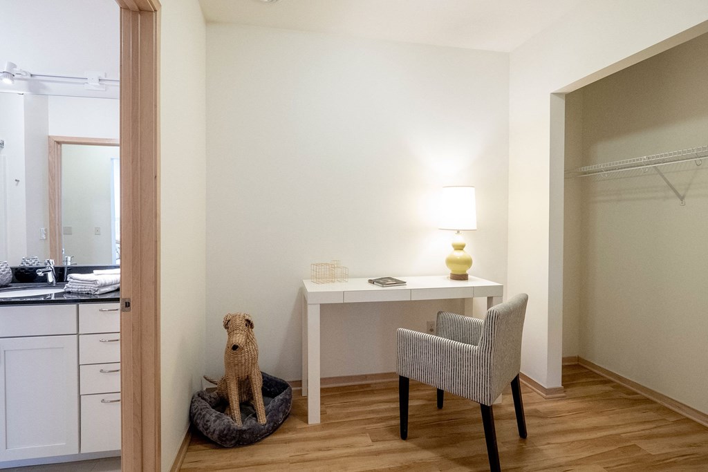A minimalist room here at 7west Apartment Homes with wooden flooring features a white desk with a lamp, a striped chair, and a woven dog in a bed. An open closet and bathroom are visible.