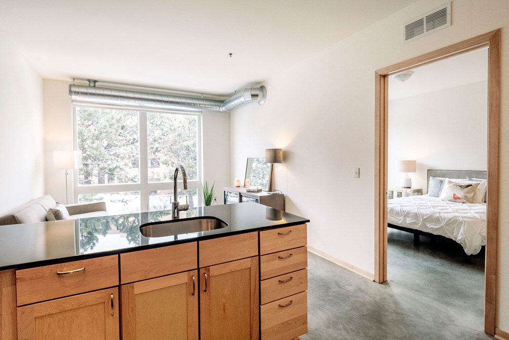 Modern apartment interior here at 7west Apartment Homes with an open kitchen, featuring wooden cabinets and a black countertop. Bright living area with a window and a glimpse of a cozy bedroom.