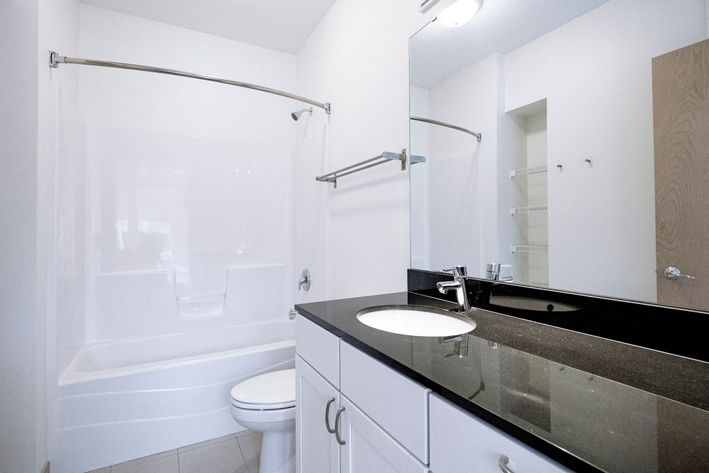 Modern bathroom here at 7west Apartment Homes with white walls and fittings. Features a bathtub with a shower curtain, a toilet, and a sleek black countertop with a sink.