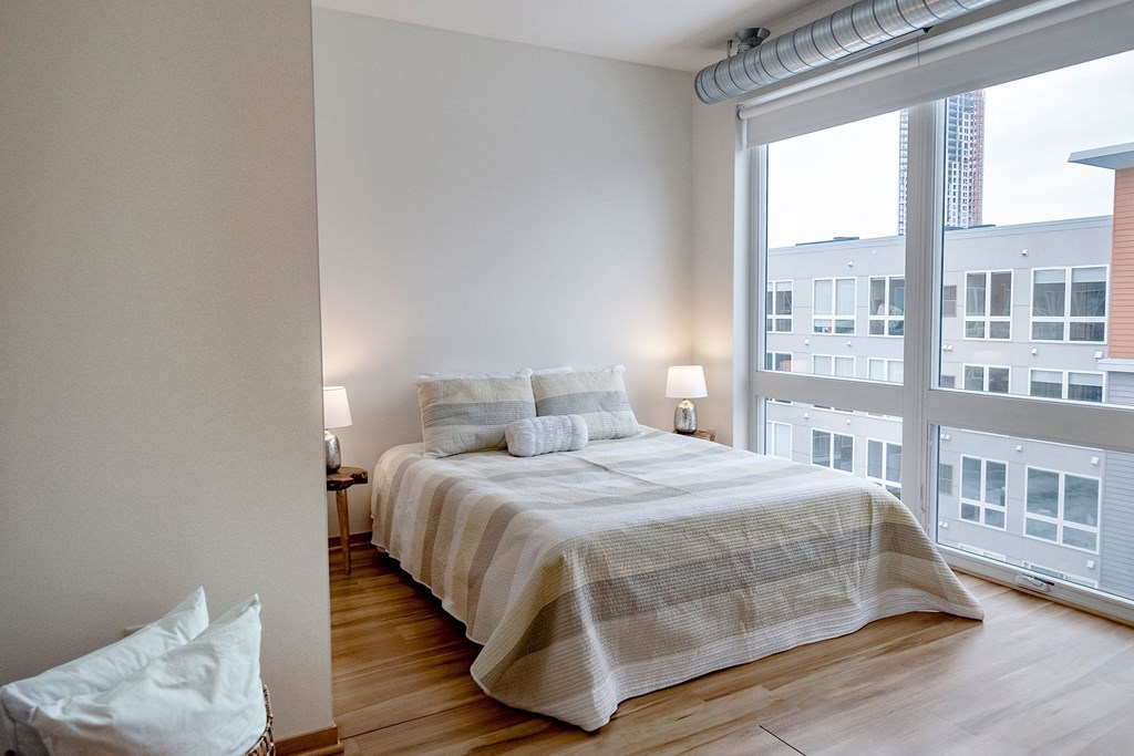A modern bedroom here at 7west Apartment Homes with a neatly made bed featuring striped bedding. Two lamps on bedside tables provide soft lighting. Large windows reveal a city view.