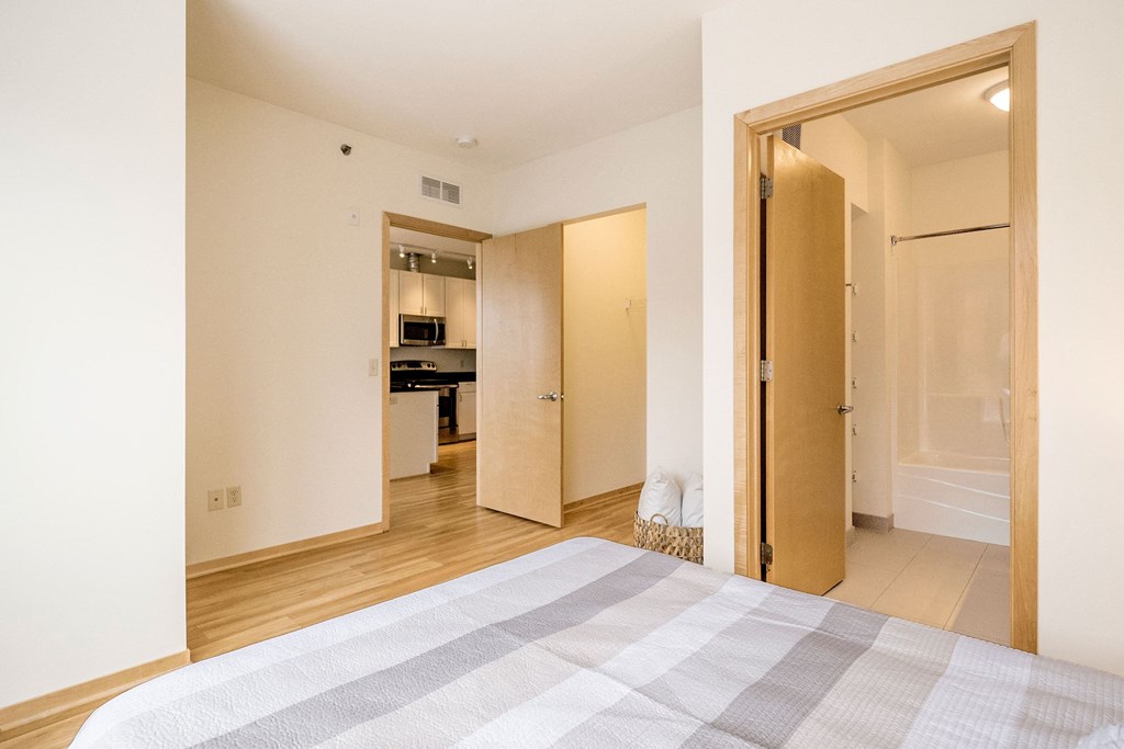 A bright bedroom here at 7west Apartment Homes featuring a neatly made bed, light wood floor, and cream walls. Open doors reveal a modern kitchen and a bathroom, creating a cozy, inviting ambiance.