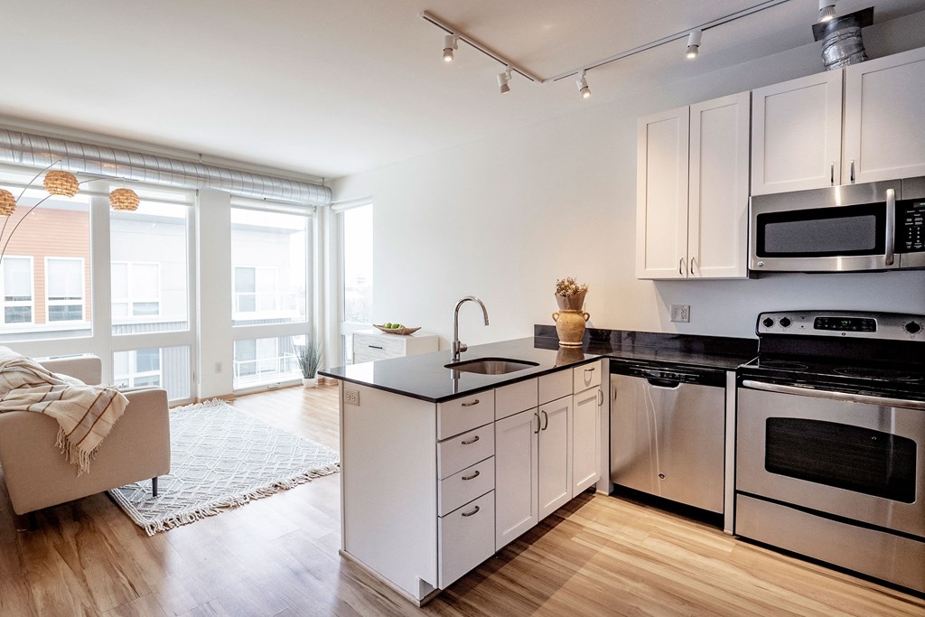 Modern apartment interior here at 7west Apartment Homes with a bright open-plan living room and kitchen, featuring large windows, light wood floors, a beige sofa, and sleek appliances.