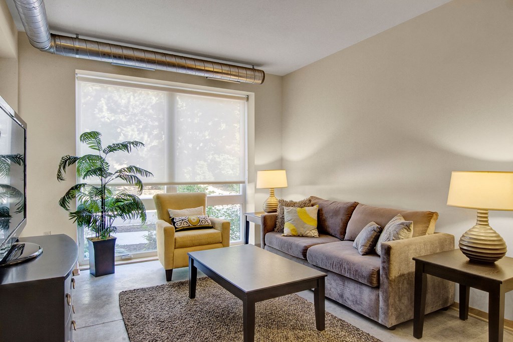 A cozy living room here at 7west Apartment Homes with a brown sofa and yellow armchair next to large windows. Warm lighting from lamps and greenery create a relaxed, inviting atmosphere.