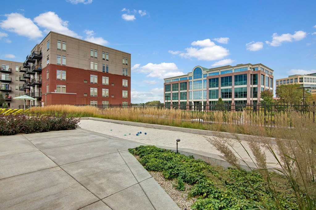 Urban scene with modern buildings here at The Covington Apartment Homes under a blue sky. A red-brick apartment and glass office face a landscaped area with grass and a bocce court.