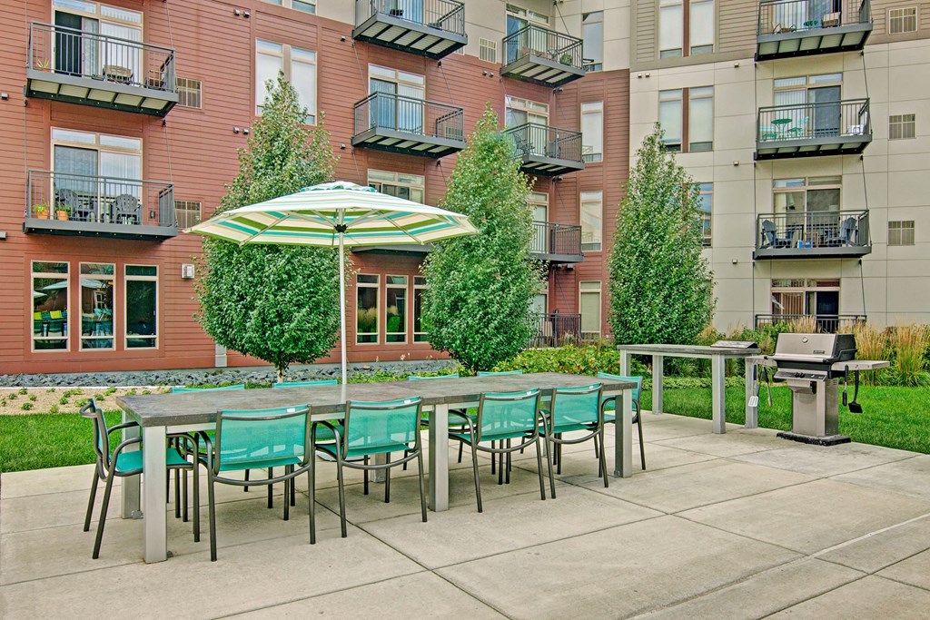 Modern The Covington Apartment Homes courtyard featuring a dining set with green chairs under a striped umbrella, surrounded by lush greenery and a grill for barbecuing.