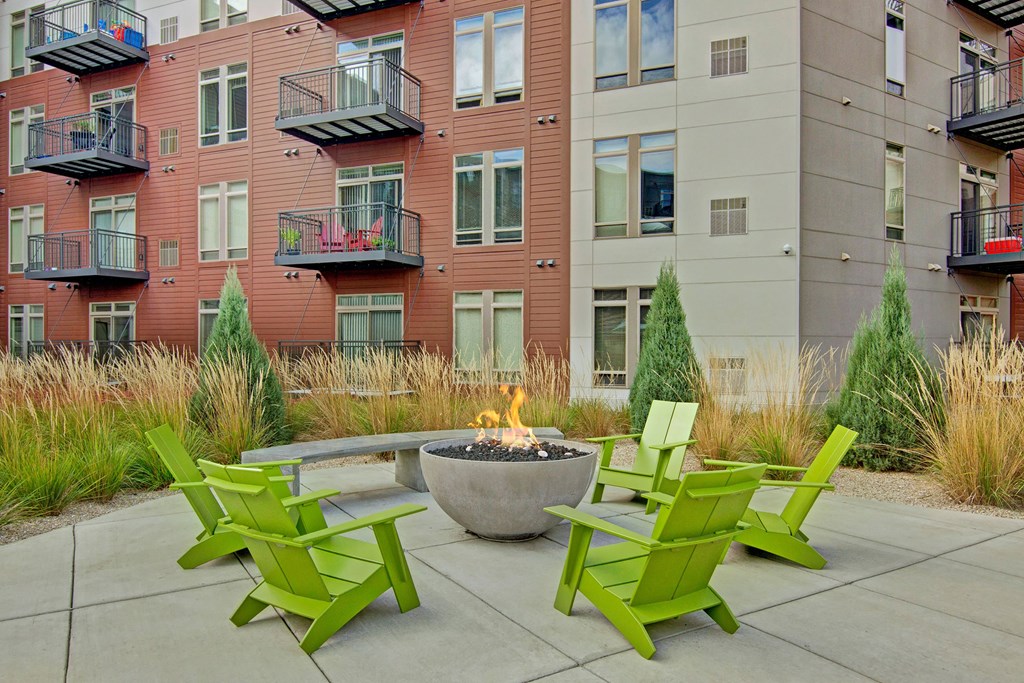 Modern The Covington Apartment Homes courtyard with green Adirondack chairs around a fire pit, surrounded by ornamental grasses and a multi-story building with balconies.