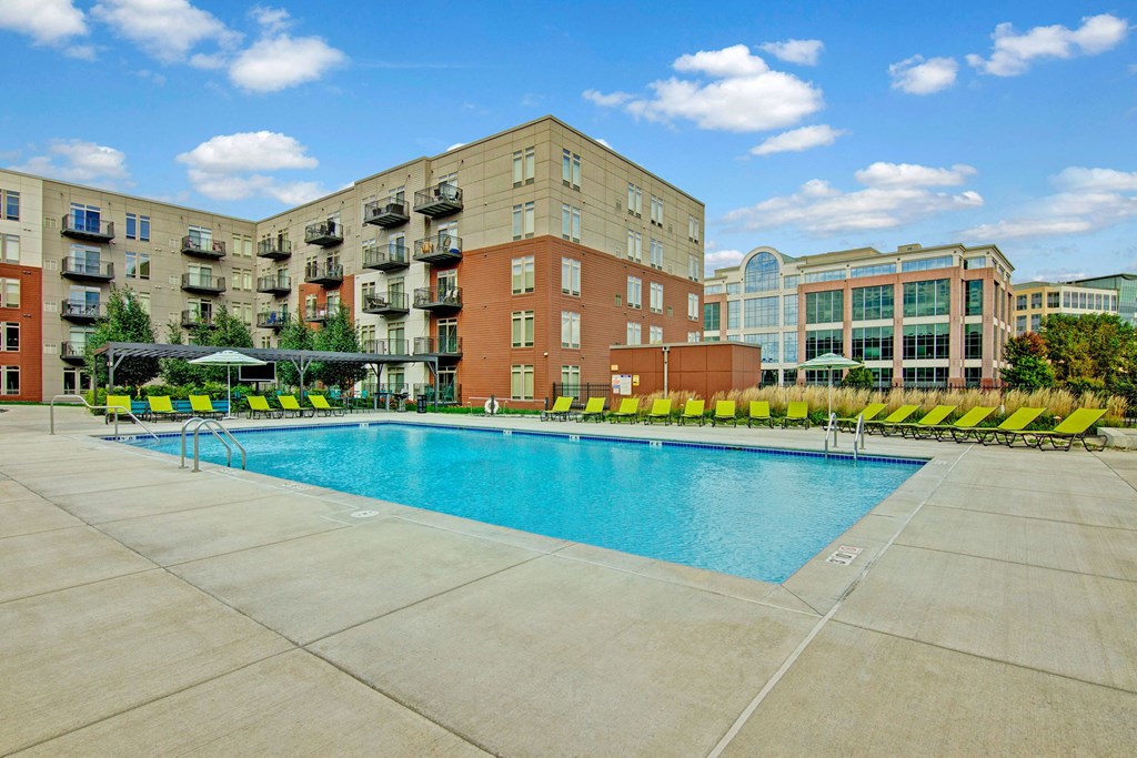 Outdoor The Covington Apartment Homes complex pool area with clear blue water and surrounding green lounge chairs. Modern buildings and a blue sky with clouds in the background.