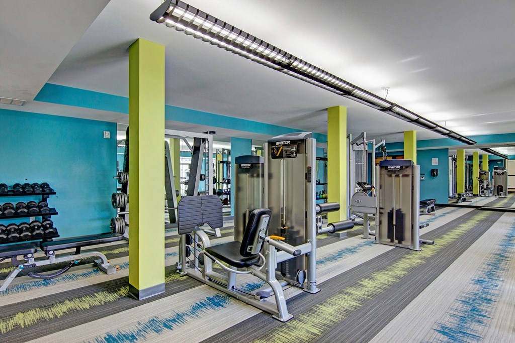 Covington Fitness Center Cardio Equipment Modern gym interior here at The Covington Apartment Homes with vibrant yellow and blue accents features a variety of exercise machines, weights, and mirrored walls, creating an energetic atmosphere.