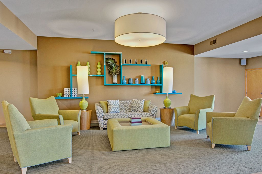 Modern lounge here at The Covington Apartment Homes with green armchairs and a patterned sofa around a square ottoman. Beige walls feature blue shelves with decorative items. Warm, inviting ambiance.