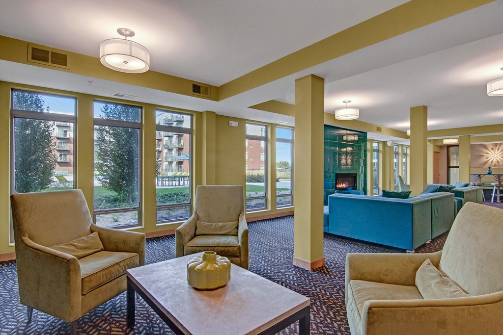 Modern lounge here at The Covington Apartment Homes with large windows and soft lighting. Features beige armchairs, a coffee table with a yellow vase, and a teal sofa by a fireplace, creating a cozy atmosphere.