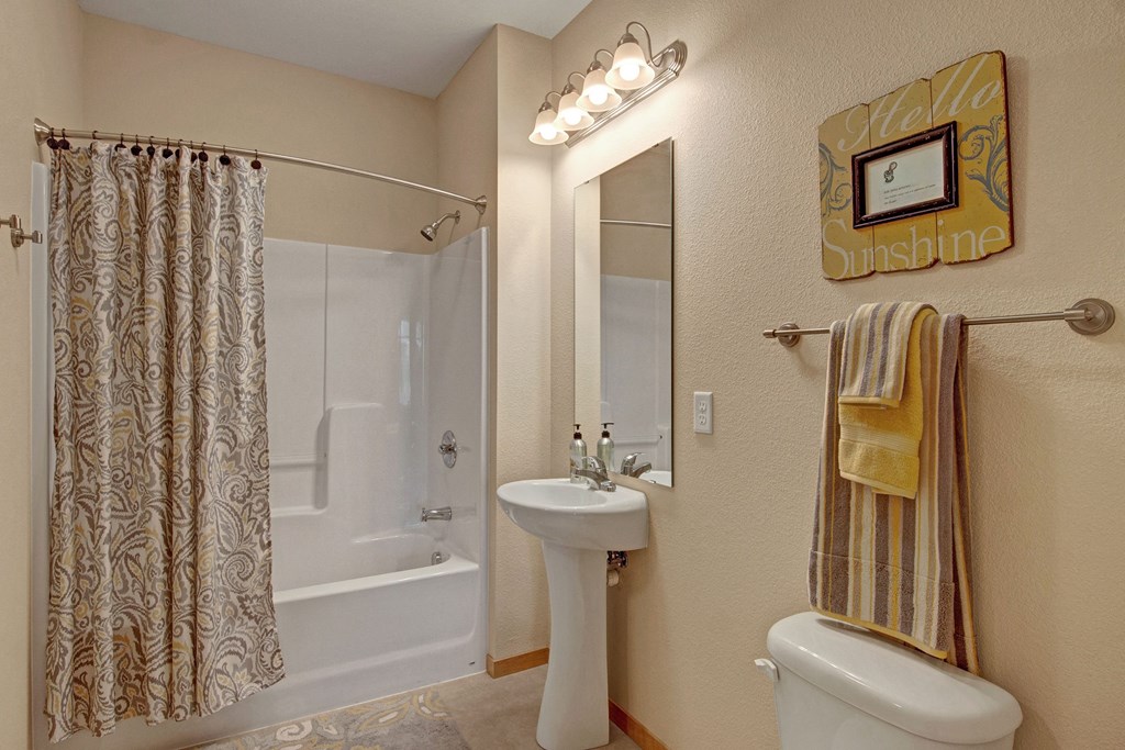 Bathroom here at The Covington Apartment Homes with beige walls features a white bathtub and a patterned shower curtain. A pedestal sink with a mirror is flanked by a towel rail and framed art saying "Hello Sunshine."