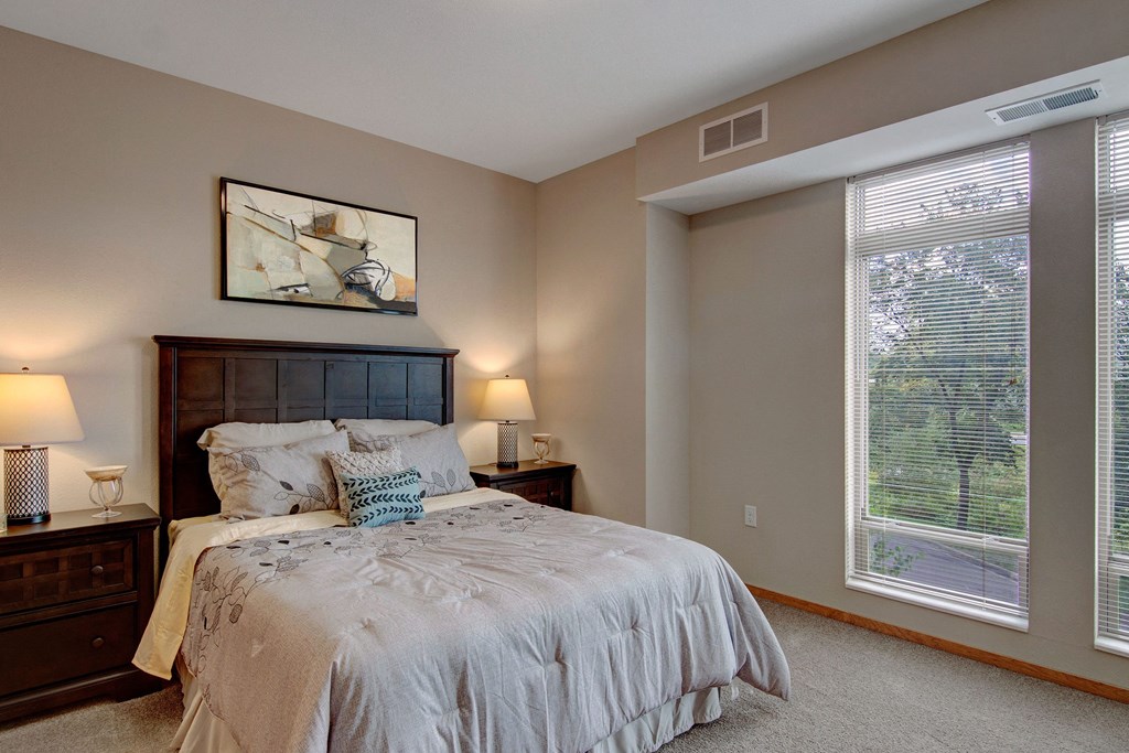 Covington Furnished Bedroom View A cozy bedroom here at The Covington Apartment Homes with a dark wood bed frame and beige bedding. Two bedside tables with lamps flank the bed. A large window with blinds lets in natural light.