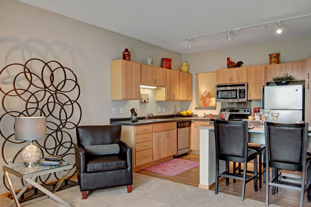 Cozy modern living space with a kitchen and seating area here at The Covington Apartment Homes. Light wood cabinets, stainless steel appliances, and sleek black chairs create a warm, inviting ambiance.