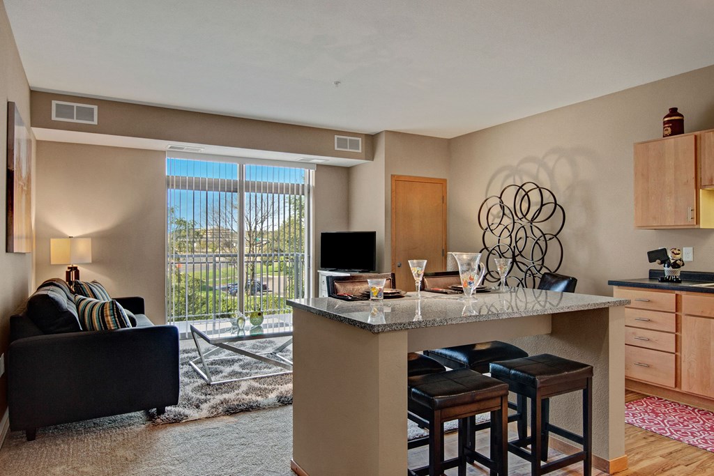 Modern living room here at The Covington Apartment Homes with beige walls, featuring a dark couch, striped pillows, and a lamp. Granite breakfast bar with stools, and intricate wall art. Light flows through large windows, creating a cozy atmosphere.