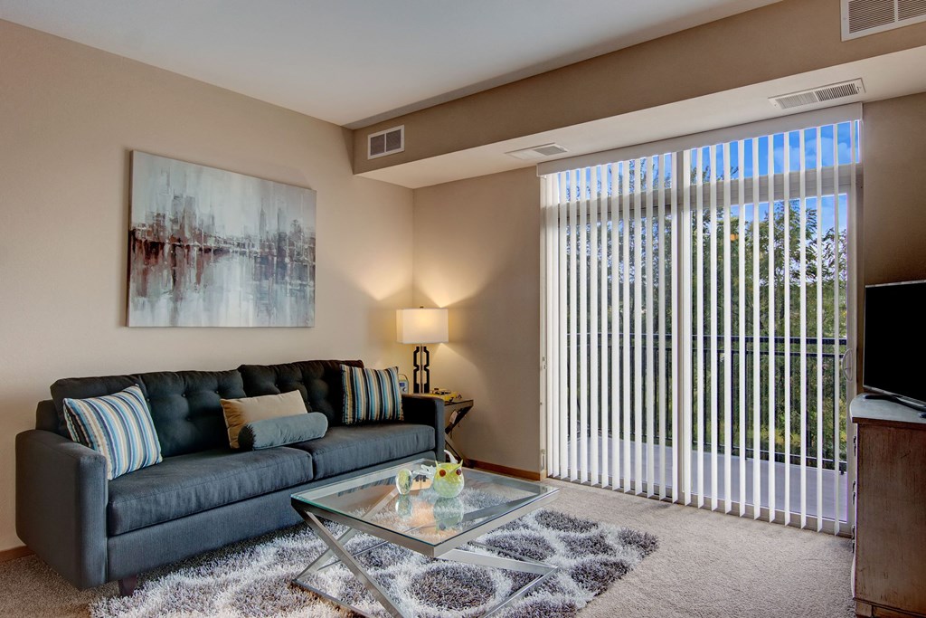 Modern living room here at The Covington Apartment Homes with a gray sofa, striped pillows, abstract wall art, and a glass coffee table. Bright light streams through vertical blinds.