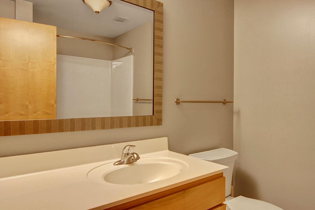 Simple bathroom here at Lofts at Farmers Market Apartment Homes with beige walls, wooden cabinet, and large mirror. White sink, metal faucet, toilet, and towel bar create a clean, minimal look.