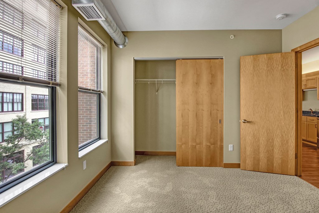 Empty bedroom here at Lofts at Farmers Market Apartment Homes with beige walls and carpet, featuring large windows with blinds, an open closet with a sliding wooden door, and a view of adjacent buildings.