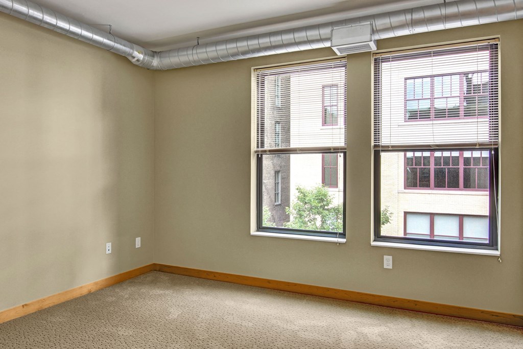 Empty bedroom here at Lofts at Farmers Market Apartment Homes with beige walls and carpet, featuring two large windows with blinds and exposed ductwork on the ceiling. Bright, neutral atmosphere.