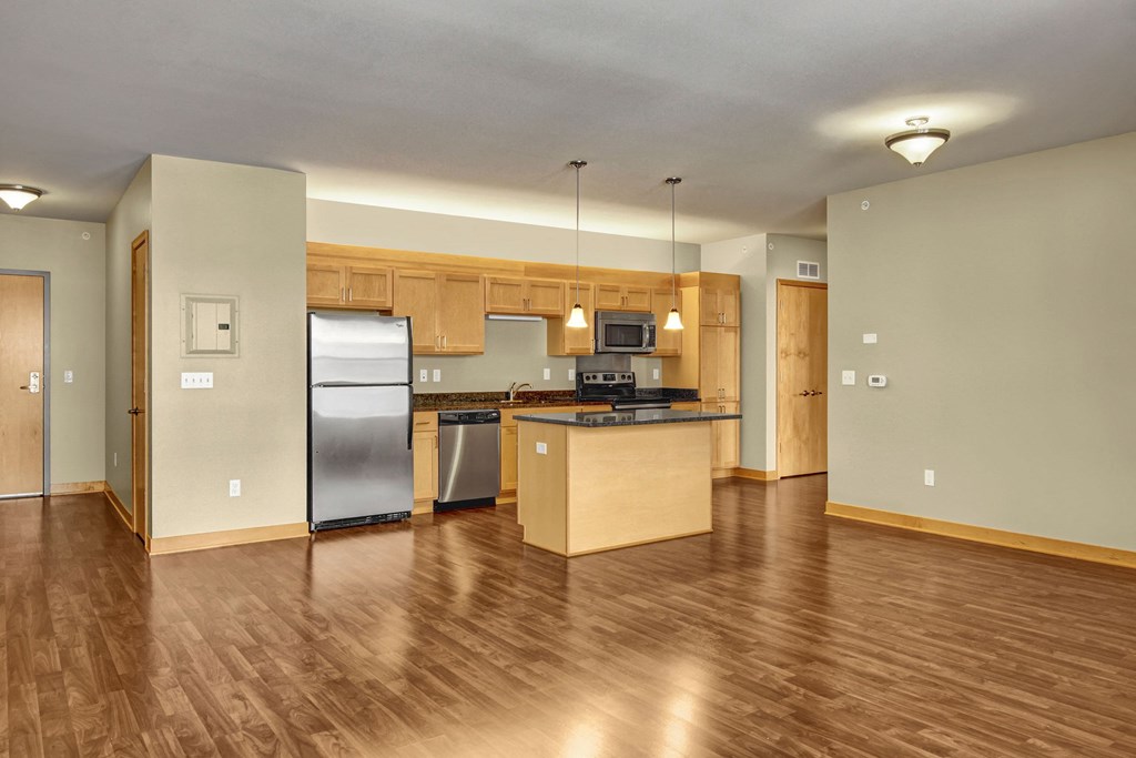 Spacious modern kitchen here at Lofts at Farmers Market Apartment Homes with wooden cabinets, island, and stainless steel appliances. Hardwood floors and pendant lighting create a warm ambiance.