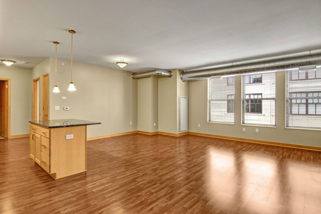 Spacious empty loft here at Lofts at Farmers Market Apartment Homes with hardwood floors, large windows, exposed ductwork, and minimalist lighting. Kitchen island with pendant lights adds warmth.