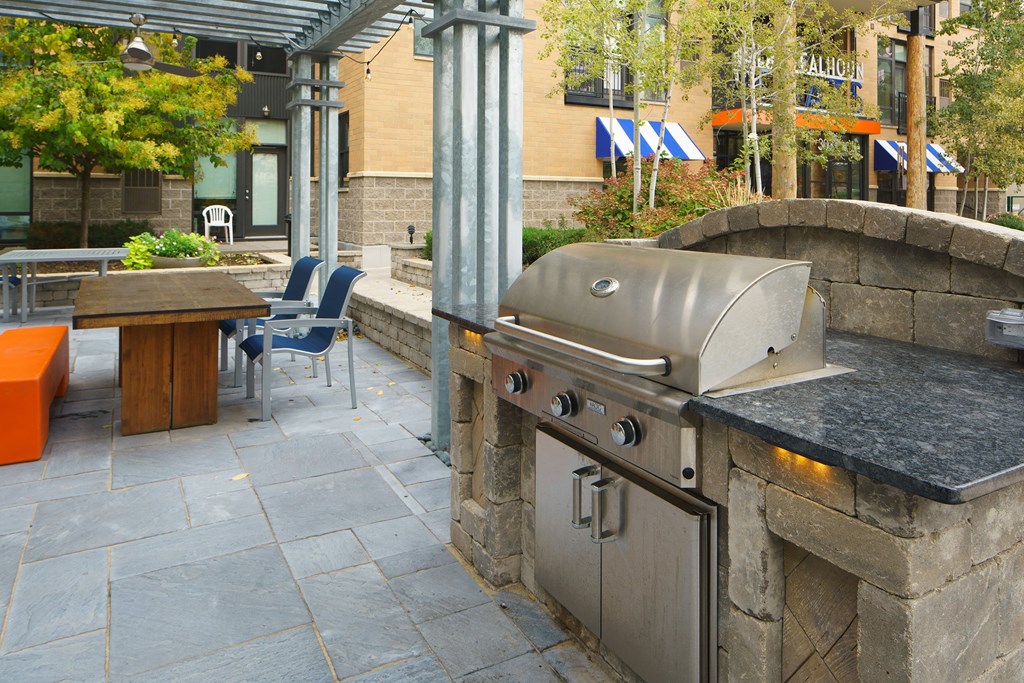 Modern outdoor patio here at Marbella on Dean Apartment Homes with a stainless steel barbecue grill, a wooden table, orange bench, and blue chairs under a pergola, surrounded by greenery.