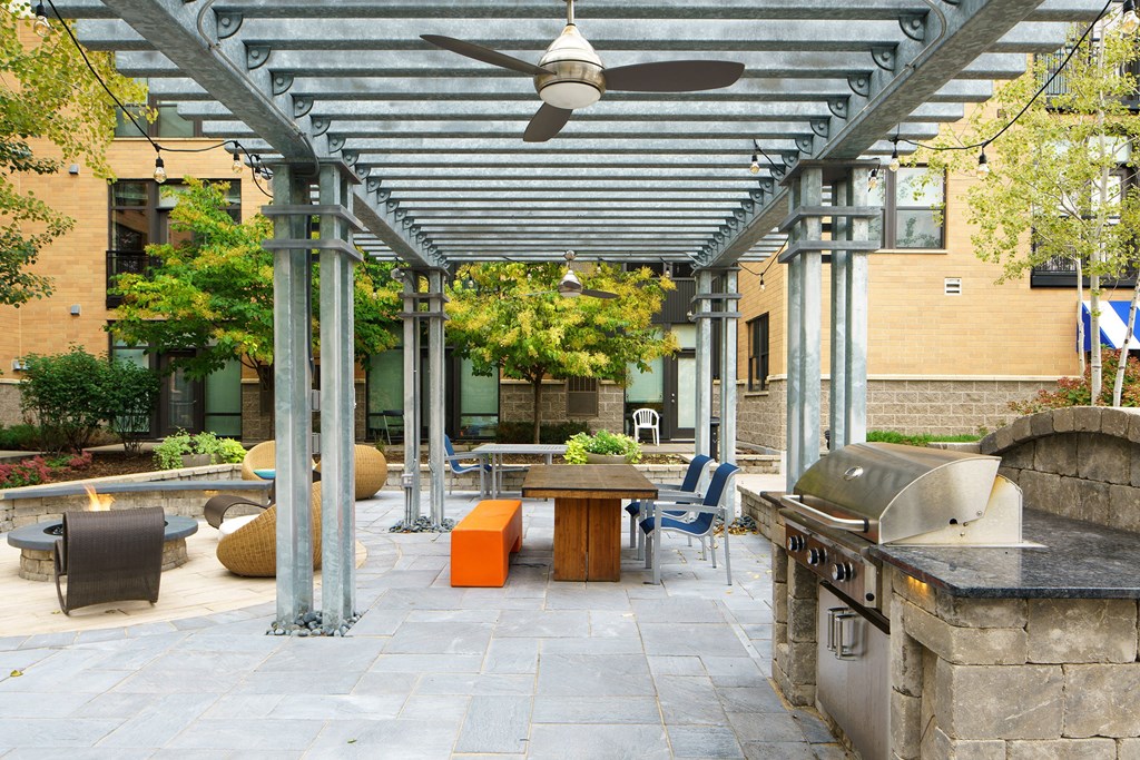 Outdoor patio with pergola, gray stone tiles, and string lights here at Marbella on Dean Apartment Homes. Features a wooden table, orange bench, barbecue grill, and fire pit. Trees add greenery.