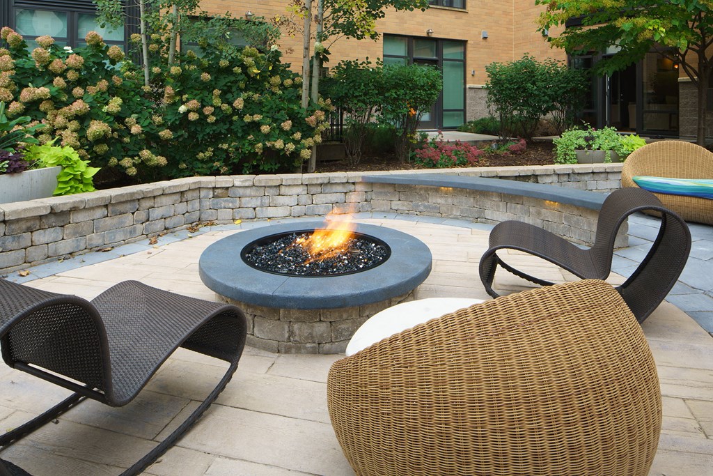 Cozy outdoor patio here at Marbella on Dean Apartment Homes with a circular fire pit, surrounded by wicker chairs. Lush greenery and a brick wall create a serene, inviting atmosphere.