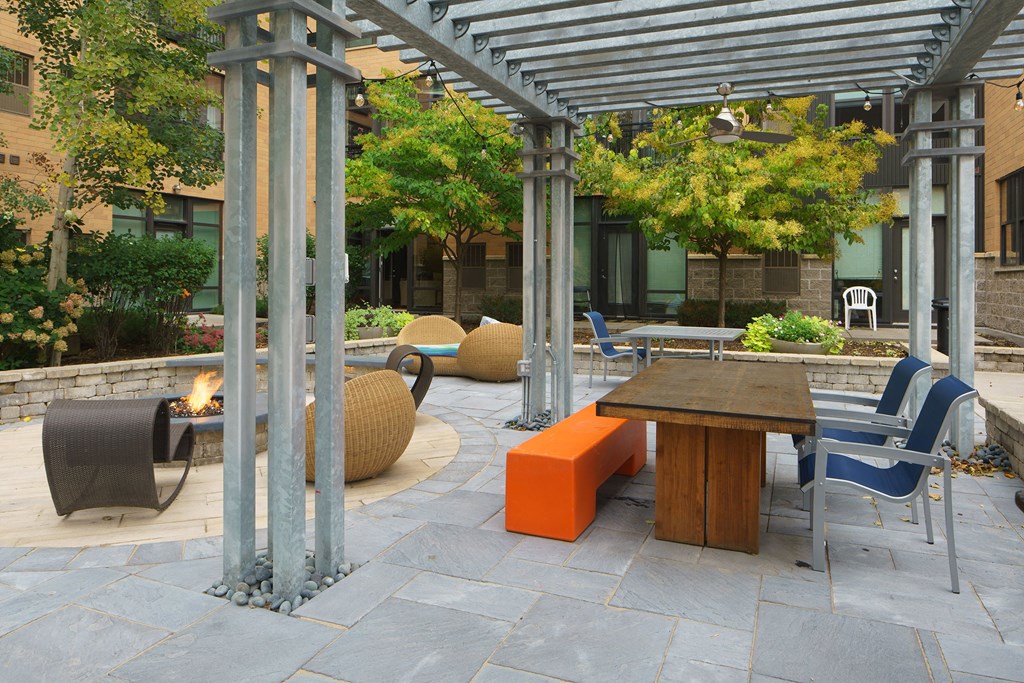 Outdoor patio here at Marbella on Dean Apartment Homes with modern metal pergola, wooden table, orange bench, and blue chairs. Woven lounge chairs surround a fire pit, all amidst lush greenery.