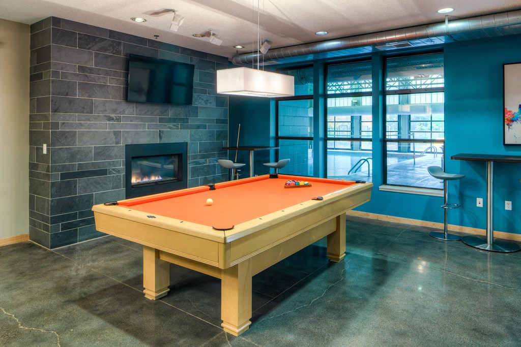 Modern game room here at Marbella on Dean Apartment Homes featuring a vibrant orange pool table on a polished floor, dark stone fireplace, wall-mounted TV, and bright windows, evoking a sleek, cozy vibe.
