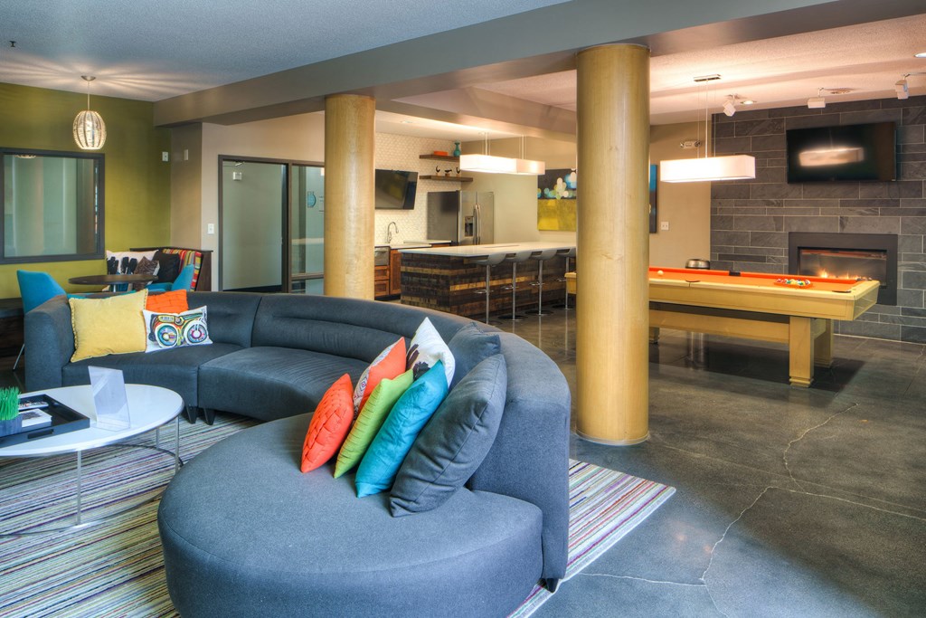 Stylish lounge here at Marbella on Dean Apartment Homes with a curved gray sofa and colorful cushions, a modern kitchen, pool table, and a cozy fireplace, evoking a relaxed, inviting vibe.