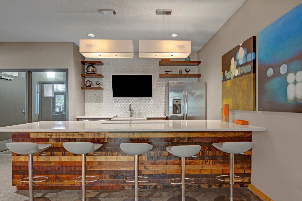Modern kitchen here at Marbella on Dean Apartment Homes with a large island, wood accents, and metal bar stools. Stainless steel fridge, abstract art, and pendant lights add style. Warm and inviting.
