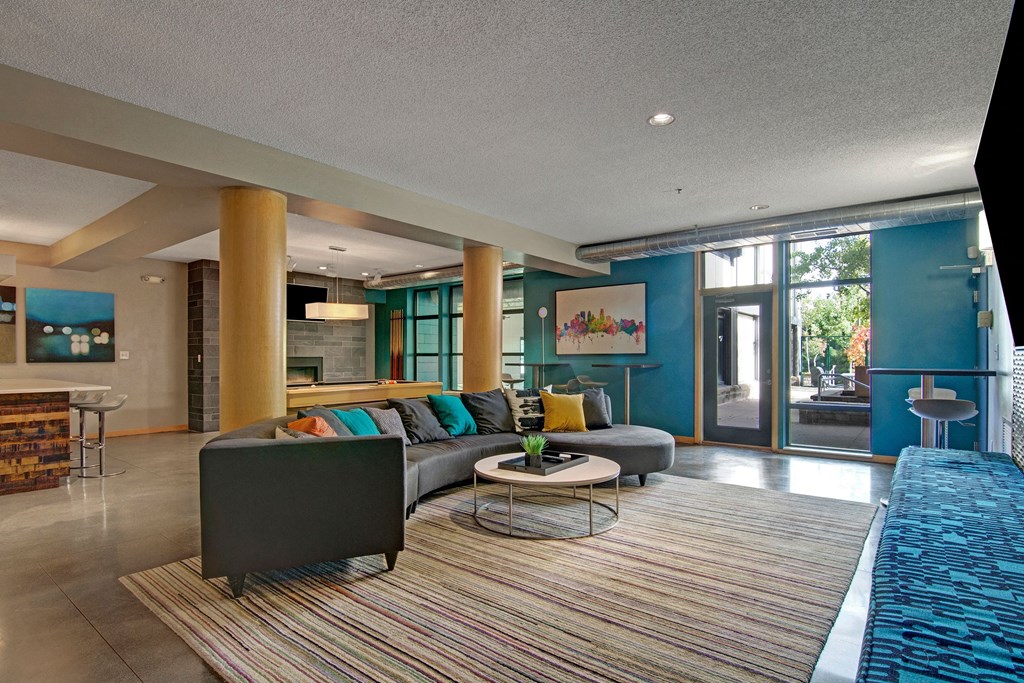 Modern living room here at Marbella on Dean Apartment Homes with a curved gray sofa adorned with colorful pillows, a round coffee table, blue walls, abstract art, large windows, and cozy ambiance.