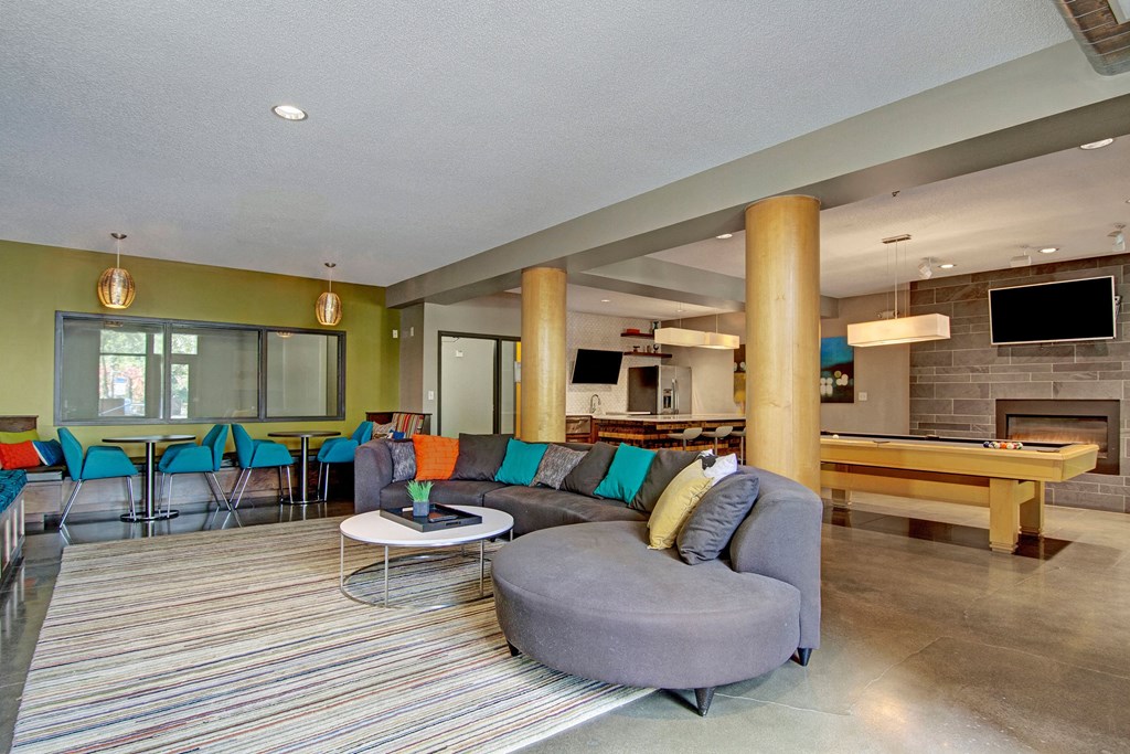 Modern communal lounge here at Marbella on Dean Apartment Homes with a curved gray sofa and colorful pillows, a striped rug, pool table, fireplace, bar seating, and large windows. Cozy ambiance.