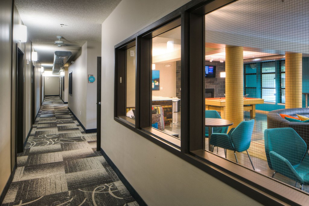 Modern hallway here at Marbella on Dean Apartment Homes with patterned carpet leads to a well-lit lounge area. Lounge features teal chairs, a pool table, and a cozy atmosphere behind glass windows.