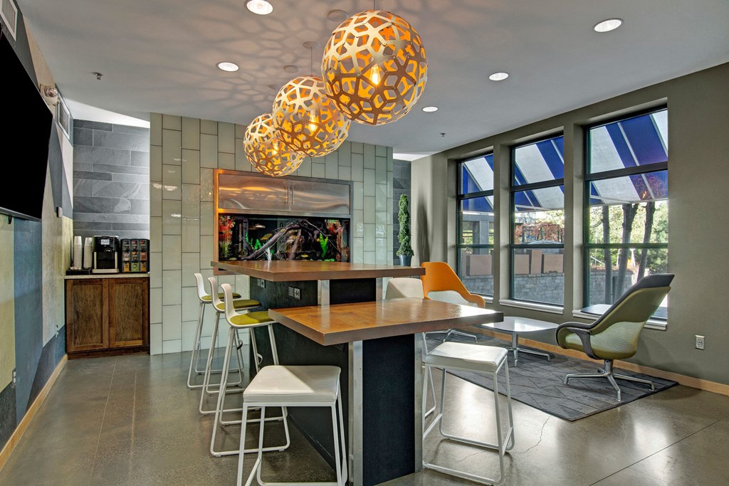 Modern resident lounge here at Marbella on Dean Apartment Homes with geometric pendant lights, a central wooden table, high chairs, and a fish tank on the wall. Large windows offer natural light.
