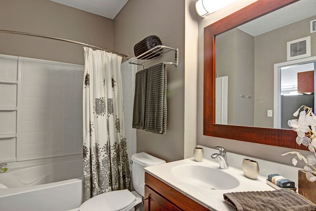 Modern bathroom here at Marbella on Dean Apartment Homes featuring a white sink with wooden cabinetry and a large mirror. Gray towels and a floral shower curtain add elegance. Soft lighting.