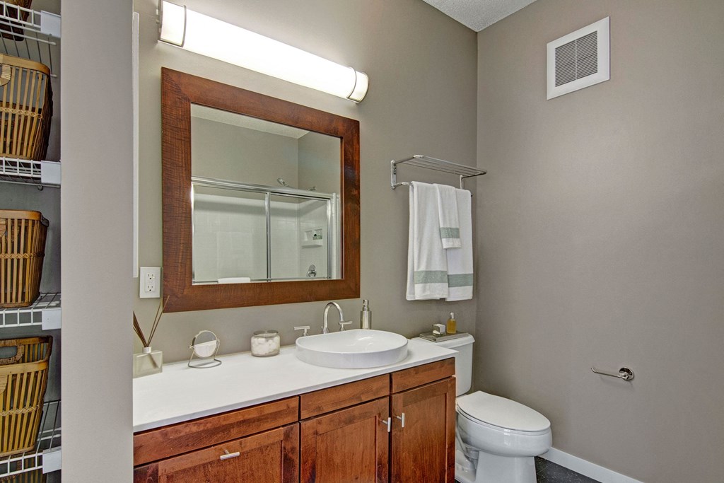 Modern bathroom here at Marbella on Dean Apartment Homes with a wooden cabinet and white countertop. Features a round sink, wall-mounted mirror, and neatly folded towels on a rack. Elegant, clean design.