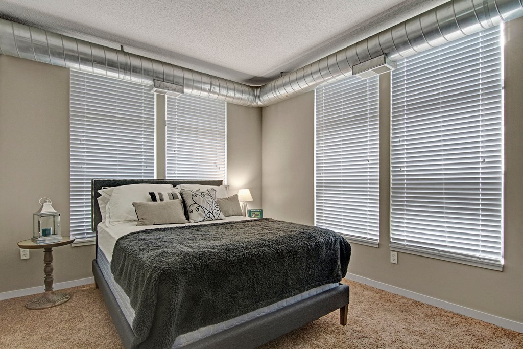 Modern bedroom here at Marbella on Dean Apartment Homes with a cozy bed, grey and white bedding, industrial ductwork, carpet, side table with a candle lantern, and blinds on large windows.