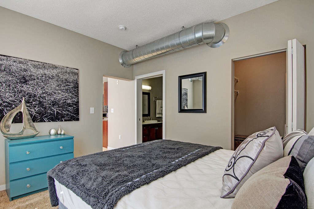 Bedroom here at Marbella on Dean Apartment Homes with beige walls features a bed with patterned pillows and a gray blanket. A blue dresser holds decor, while a silver vent adds a modern touch.