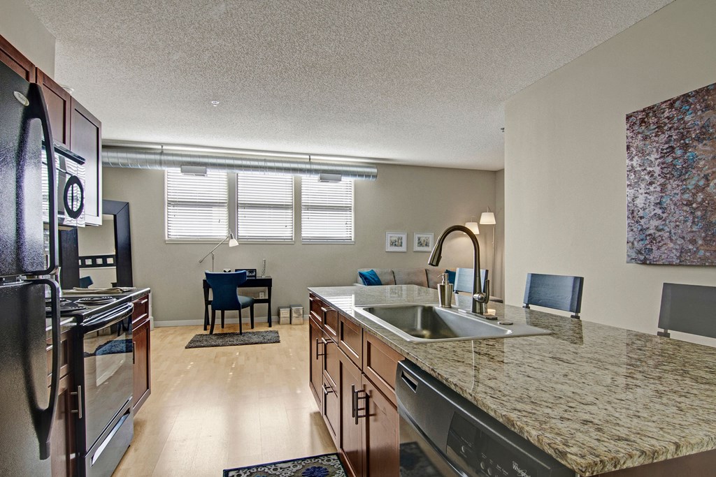 Modern apartment kitchen and living area here at Marbella on Dean Apartment Homes with granite countertops, stainless steel appliances, and a blue chair at a desk near three large windows. Cozy and inviting.