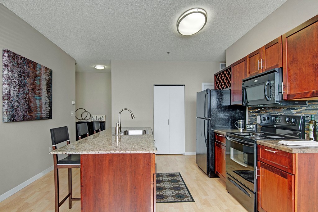 Modern kitchen here at Marbella on Dean Apartment Homes with dark wood cabinets, granite island with sink, black appliances, and colorful wall art. Cozy and welcoming ambiance.