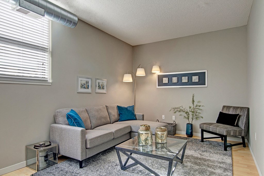 Modern living room here at Marbella on Dean Apartment Homes with gray sofa, blue and gray pillows, and glass coffee table. Decorative wall art, tall floor lamp, and indoor plant accent the space.