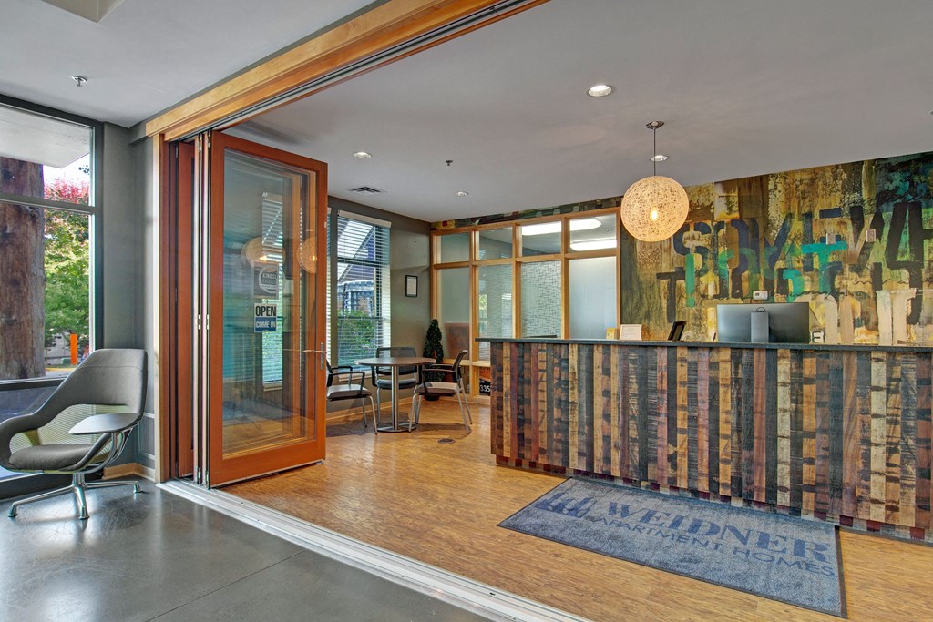 Modern office lobby here at Marbella on Dean Apartment Homes with a wood reception desk, artistic wall, round light fixture, and glass doors opening to a cozy seating area. Welcoming atmosphere.
