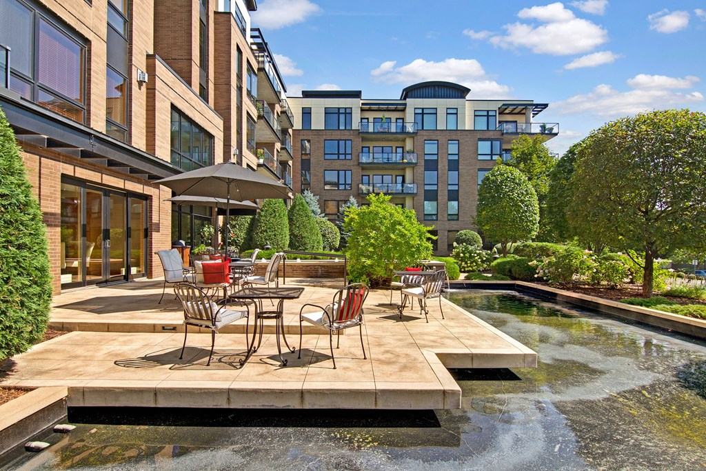 A sunny courtyard here at The Mist on Lake Minnetonka Apartment Homes features elegant wrought iron tables and chairs with red cushions, surrounded by lush greenery, near a modern brick building facade.