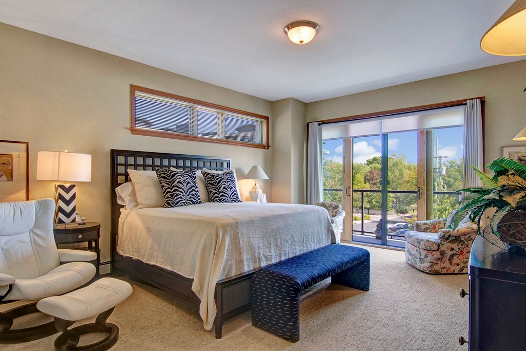 A cozy bedroom here at The Mist on Lake Minnetonka Apartment Homes features a large bed with white linens and patterned pillows. A sliding glass door reveals a bright view of lush trees, enhancing the serene ambiance.