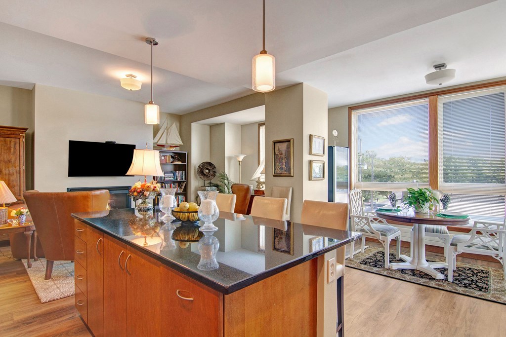 Bright kitchen and dining area here at The Mist on Lake Minnetonka Apartment Homes with a polished wooden island, pendant lights, and large windows. Includes a cozy seating area and decorative plants.