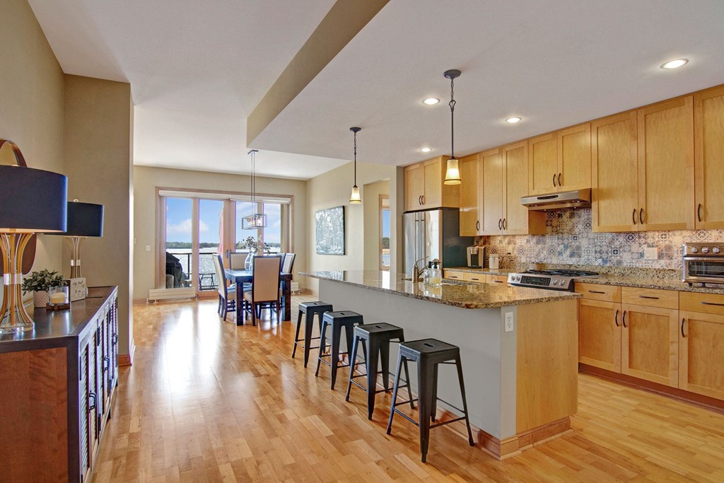 Spacious kitchen here at The Mist on Lake Minnetonka Apartment Homes with light wood cabinets, granite countertops, and a central island with stools. Dining area with a view of the lake through large windows.