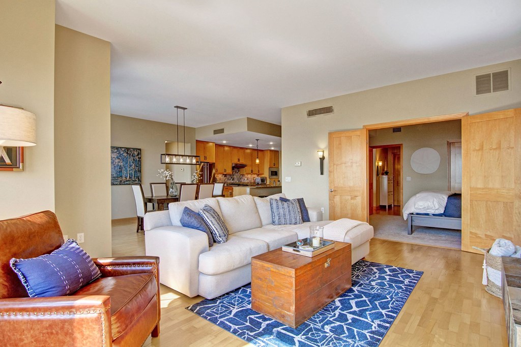 A cozy living room here at The Mist on Lake Minnetonka Apartment Homes with a brown leather chair, a beige sofa adorned with blue pillows, and a wooden coffee table on a blue patterned rug. Open space leads to a dining area and kitchen with warm lighting.