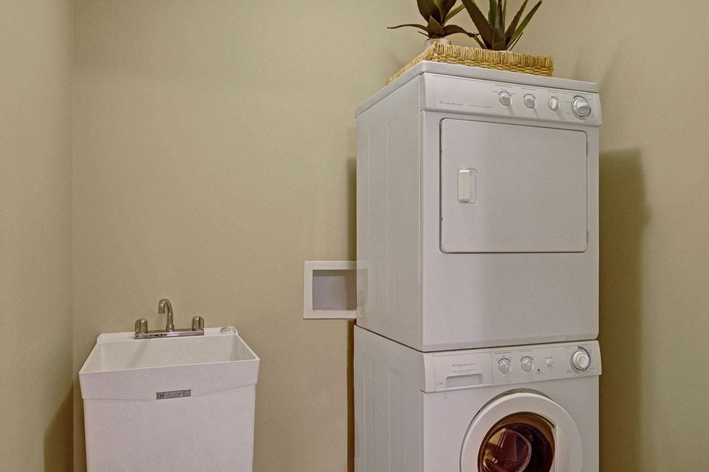 Stacked white washer and dryer in a laundry room beside a utility sink here at The Mist on Lake Minnetonka Apartment Homes. A potted plant decorates the top of the dryer, adding a touch of greenery.